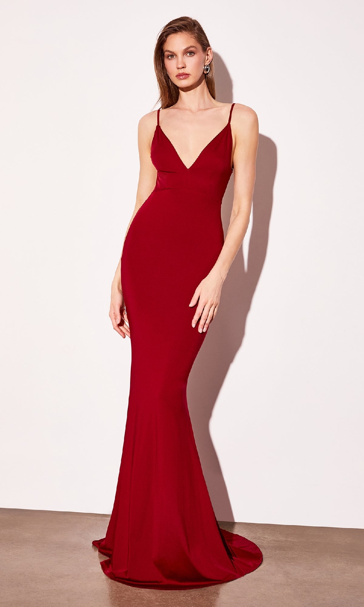 A woman stands against a plain white wall wearing the Astee T905 Open-Back Long Bodycon Prom Dress, a sleeveless, fitted red gown with thin straps and a V-neckline.